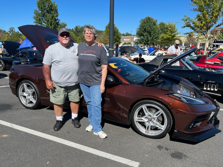 Two people standing beside a brown C8 Corvette convertible.