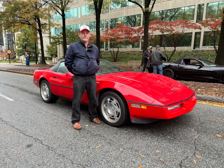 A man wearing a jacket is standing beside a Corvette convertible.