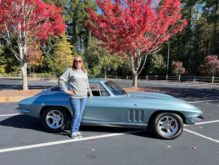 A woman is standing beside a second-generation light blue Corvette coupe.