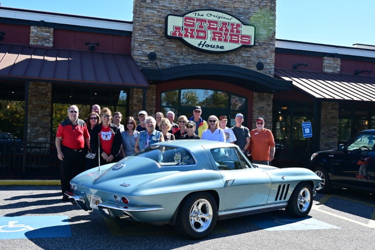 A crowd of people standing outside a restaurant beside a classic Corvette