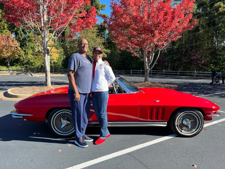 Two people are standing beside a C2 second-generation Corvette convertible in a parking lot.