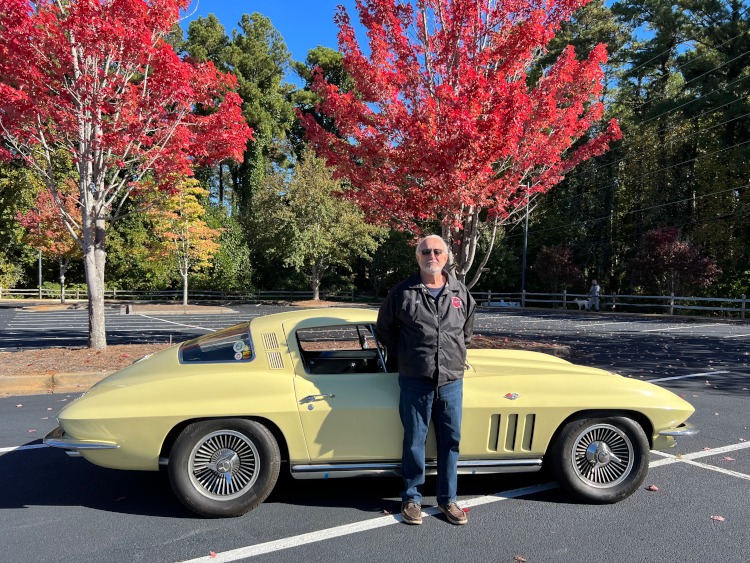 A man is standing beside a yellow second-generation Corvette coupe.