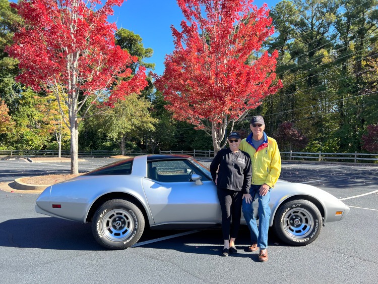 Two people are standing beside a silver Corvette coupe.
