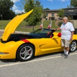 A man is standing beside a yellow C5 Corvette convertible.