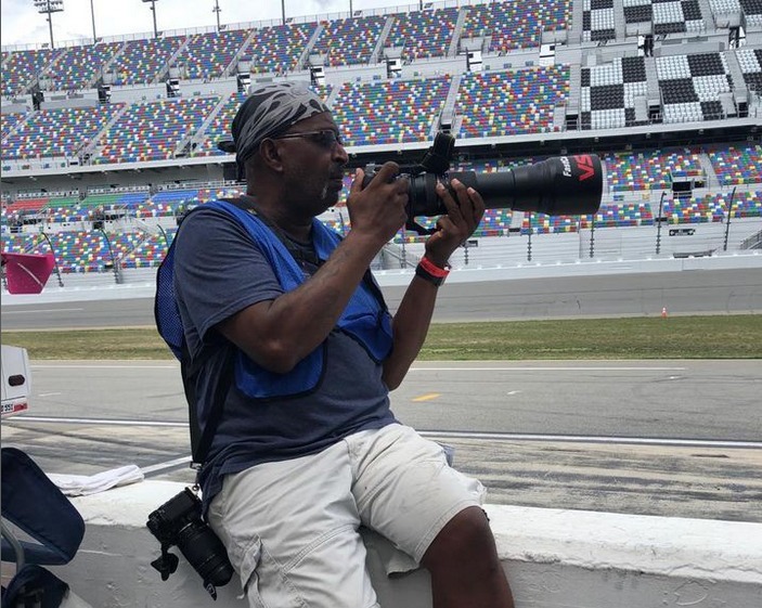 A man holding a camera sitting on a race barrier.