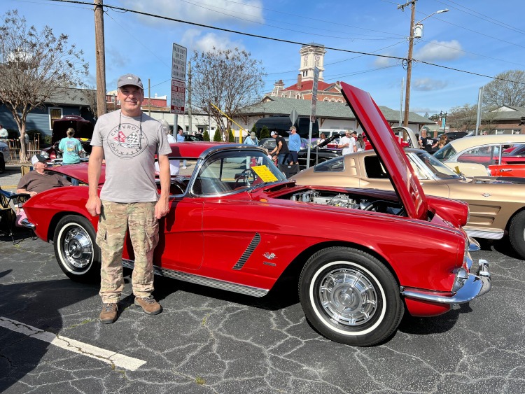 A man is standing beside a red 1962 roadster.