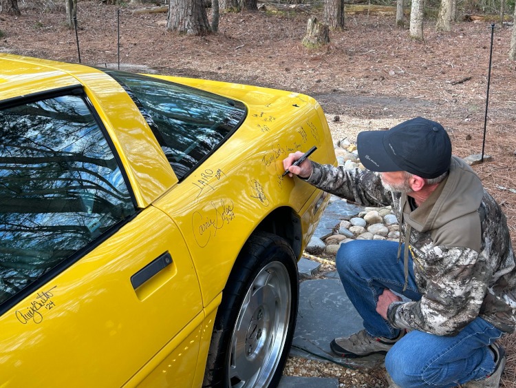 A man is signing the back fender of a yellow Corvette.