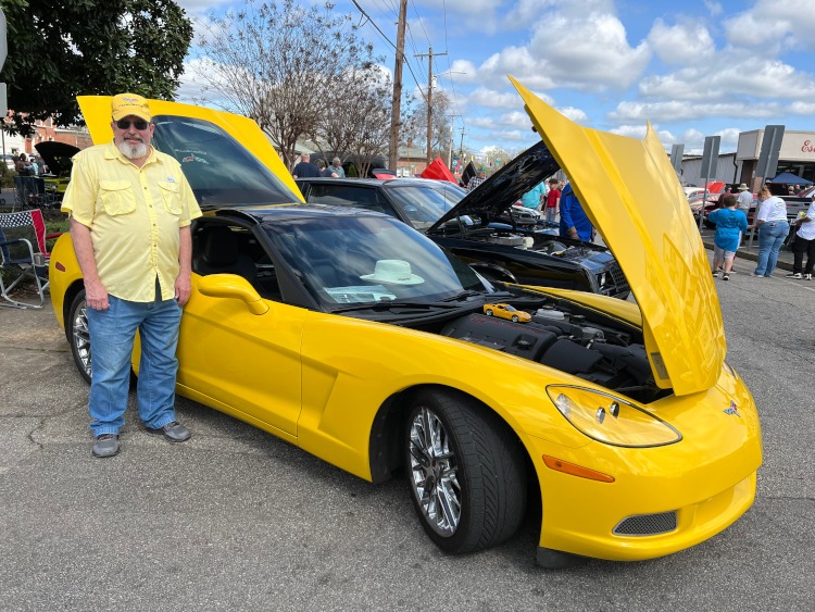 A man standing beside a 2013 C6 Corvette coupe at a car show.