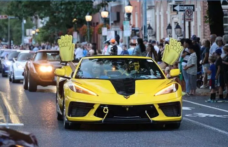 Yellow C8 Corvette convertible with a person wearing a 'Chevy' themed mask and two giant foam hands waving, leading the Downtown Carlisle Corvette Parade during Corvettes at Carlisle 2025. Photo from Vettes of Atlanta Magazine.