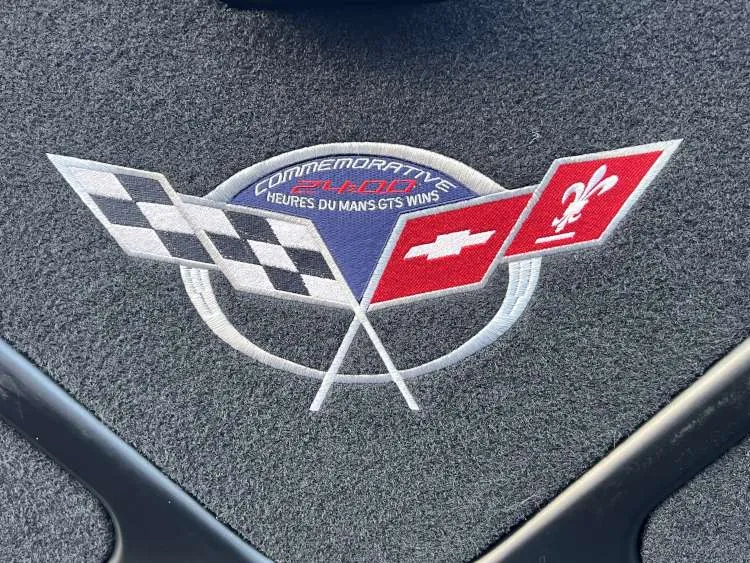 Close-up view of the rear cargo area carpeting in a 2004 Chevrolet Corvette Z06, featuring the high-contrast "Z06" logo embroidery. The red "Z" and silver "06" numbering are stitched into the black automotive-grade carpet, showcasing the specific branding of the Fixed Roof Coupe performance model.