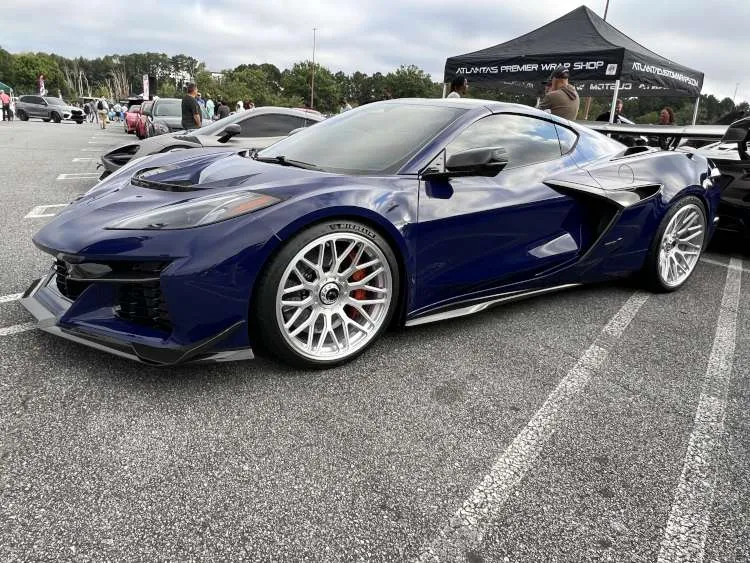 Full profile view of a Hysteria Purple 2025 Corvette ZR1 parked at an outdoor event, showcasing its long-tail silhouette, carbon fiber ground effects, and silver 20-spoke forged aluminum wheels.