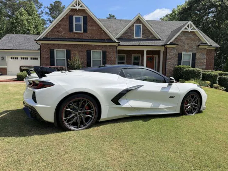 A full side-profile view of a white 2023 C8 Corvette 70th Anniversary edition convertible parked on a green lawn. The car features a black retractable hardtop, distinctive multi-spoke dark alloy wheels with a red accent stripe, and a prominent rear wing spoiler. A large, two-story brick and stone suburban house serves as the background under a clear blue sky.